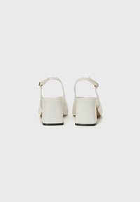 White heeled sandals with an open back, featuring a wide heel, ankle strap, and smooth glossy finish. Black accents on the sole.