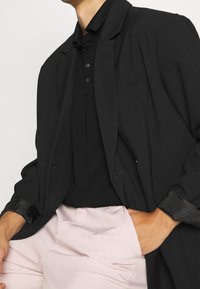 Svart kavaj över en svart polo med en slät yta. Ljusrosa byxor med fickor. Snygg, skräddarsydd design och subtila detaljer.