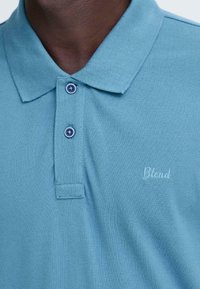 Polo bleu clair en coton texturé, avec une patte de boutonnage à trois boutons et un logo brodé "Blend" de couleur assortie.