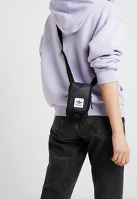 Borsa a tracolla nera con un patch logo frontale, forma rettangolare, tracolla regolabile, indossata su una felpa leggera viola e jeans scuri.