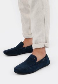 Scarpe slip-on in suede blu navy con punta arrotondata, cuciture decorative e dettagli metallici. Indossate con pantaloni di colore chiaro arrotolati.