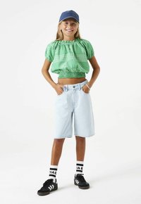 Top corto verde con mangas abullonadas y patrones blancos, pantalones cortos de mezclilla azul claro, zapatillas negras y calcetines con rayas blancas y negras.