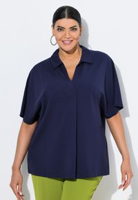 Navyblauw oversized blouse met een kraag, korte mouwen en een voornaaddetail, gedragen met felgroene pants.