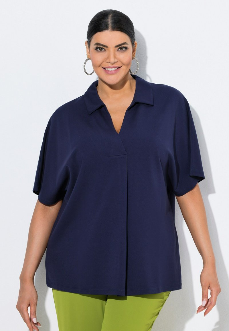 Navyblauw oversized blouse met een kraag, korte mouwen en een voornaaddetail, gedragen met felgroene pants.