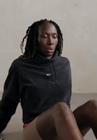 Sort hættetrøje med Nike-logo, siddende på en glat overflade, viser brun hud og tekstureret, fletningshår.