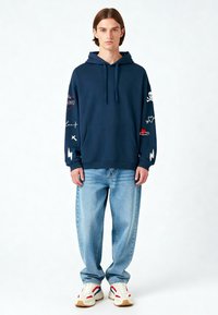 Felpa con cappuccio blu navy, dotata di tasca frontale, con design e grafiche ricamate colorate sulle maniche. Indossata con jeans azzurri.