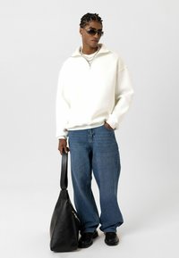 Witte fleece pullover met een ritssluiting bij de kraag, blauwe jeans, zwarte leren tas en zwarte schoenen. Model draagt een zonnebril en accessoires.