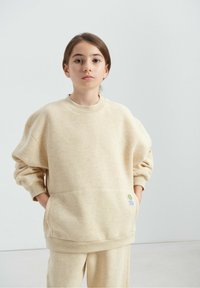 Sweat-shirt oversize beige clair à manches longues, col rond côtelé et poche avant avec un petit patch logo. Texture de tissu douce.