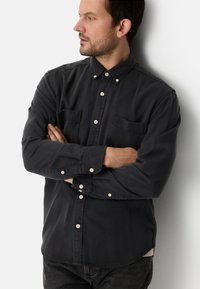 Chemise noire boutonnée avec deux poches avant, manches longues et boutons blancs. Le tissu semble lisse avec un léger éclat.