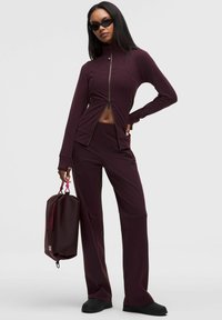 Bordowa sportowa kurtka z zamkiem błyskawicznym, długimi rękawami i dopasowanymi spodniami. Gładka tkanina, subtelne detale w szwie. Czarne buty i fakturowana bordowa torba.