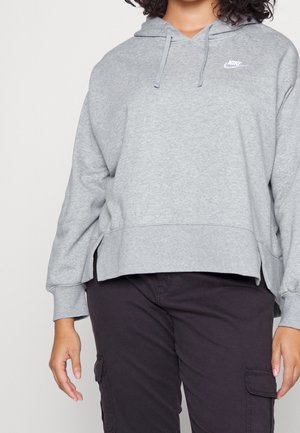 Osoba w jasno szarej bluzie z kapturem Nike ze sznurkami, zestawiona z czarnymi spodniami typu cargo na jednolitym tle.