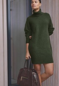Grüne Strick-Turtleneck-Kleid mit geripptem Saum und Bündchen, kombiniert mit einer dunkelbraunen Wildleder-Handtasche mit einem Reißverschlussfach und Ledergriffen.