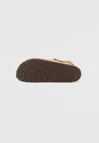 Ruskea kumipohja, jossa on aaltoileva kuvio; beige yläosa kultaisilla solkikoruilla ja teksturoiduilla hihnoilla. Pohjalla on monimutkaisia kuvioita.