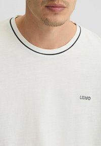 Valge puuvillane T-särkk, ümmarguse kaeluse ja musta äärega, vasakus rinnaosas on väike sinine logos "LIUJO". Pehme tekstuur.