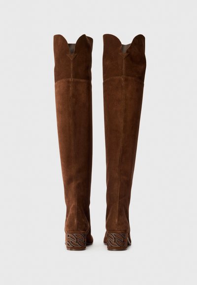 Botas de ante marrón hasta la rodilla con borde superior en forma de escalloped y tacón decorativo tallado. Textura suave y diseño ajustado para contornear la pierna.