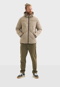 Chaqueta de plumas beige con diseño acolchado y cremallera negra, que cuenta con una capucha y dos bolsillos laterales; combinada con pantalones verde oliva y zapatillas negras.