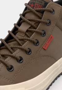 Brown synthetic sneaker with textured leather, black eyelets, and brown laces. Features red "WATER REPELLENT" tag and cream-colored sole.