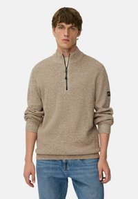 Jonge man in een beige gebreide trui met een ritssluiting tot de helft en blauwe jeans, staand met zijn handen ontspannen langs zijn zij tegen een effen achtergrond.
