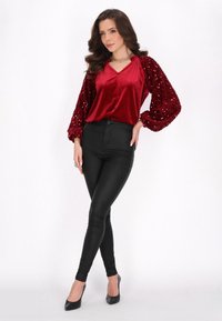 Bluză din catifea roșie cu mâneci bufante din paiete, asortată cu pantaloni skinny negri și pantofi cu toc negri. Bluza are un decolteu în formă de V și un închizător cu nasturi.