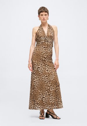 Personne portant une robe maxi sans manches à col halter imprimée léopard avec une taille plissée, des talons noirs à brides et des boucles d’oreilles créoles dorées, debout devant un fond blanc.