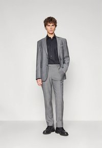 Grauer Anzug mit Karomuster und schwarzem Hemd, figurbetonte Passform, einreihiges Sakko mit einem Knopf, Hose mit flacher Vorderseite und schwarze Schnürschuhe.