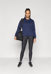 Σκούρο μπλε φούτερ, μαύρες κολάν με σχέδια και μαύρα αθλητικά παπούτσια. Το μοντέλο κρατά μια μαύρη τσάντα με λουρί, στέκεται μπροστά σε ένα μονόχρωμο φόντο.