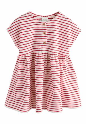 Ragazza sorridente in un vestito a righe rosse raccoglie un'arancia da un rigoglioso albero di agrumi in un soleggiato giardino all'aperto.