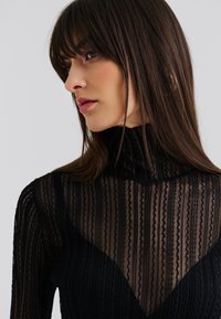 Svart mesh-top med vertikale stripete mønstre, høy hals og lange ermer. Delikate teksturer fremhever det gjennomskinnelige designet.