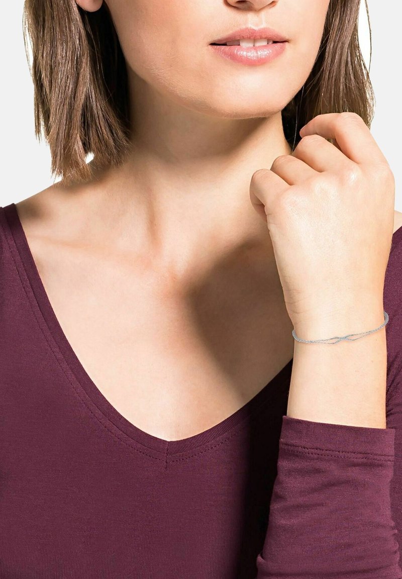 Silverfärgad armband med en sammanflätad design, bärs på en handled. Bäraren har en vinröd V-ringad topp och kort hår, med fokus på accessoaren.