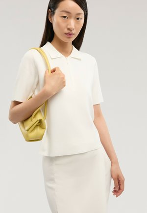 Femme portant une tenue blanche à manches courtes avec col et jupe, tenant un petit sac à main jaune sur l'épaule, fond neutre.