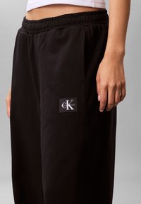 Személy fekete Calvin Klein melegítőnadrágot visel gumis derékrésszel és oldalzsebekkel, részben látható fehér rövid felsőt.