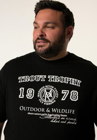 Černé bavlněné tričko s bílým grafickým textem: "TROFEJ TROUT 1978" a "VENKOVNÍ A DIVOKÁ PŘÍRODA," se zaobleným logem a dekorativními prvky.