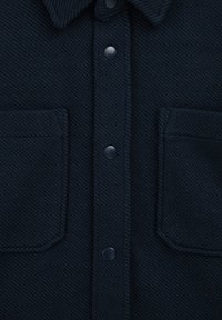 Temno modra srajca z teksturirano, tkanino. Ima usnjeni ovratnik, dva sprednja žepa in okrogle plastične gumbe.