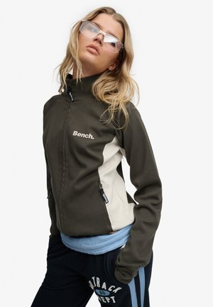 Ung kvinde iført store klare briller, mørkegrøn og beige lynlåstrøje med Bench-logo samt sorte bukser med blå striber og tekst.