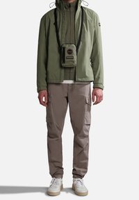 Uomo che indossa una giacca verde oliva, un maglione con zip abbinato, pantaloni cargo beige, sneakers bianche e una piccola borsa a tracolla verde oliva.