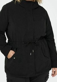 Svart jacka med dragsko i midjan, hög krage, framfickor och ett mjukt, texturerat tyg. Har silverfärgade detaljer.