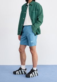 Grön denim skjorta med knappar över en vit t-shirt, i kombination med blå Nike-shorts och svarta sneakers med en vit "AIR"-logotyp.