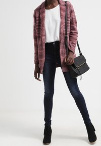 Žena v růžovém texturovaném saku, bílé blůze, tmavých skinny džínách, černých kotníkových botách a s černou crossbody taškou.
