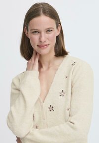 Cardigan color crema con scollo a V, texture morbida, decorato con piccoli ricami floreali in rosa e verde, e bottoni frontali.