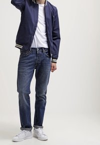 Blouson aviateur marine avec poignets rayés, t-shirt blanc, jean bleu et baskets blanches. Tenue décontractée avec une coupe ample et une texture en denim.