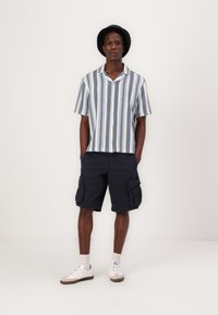 Camisa de manga corta a rayas con rayas azules y blancas, pantalones cortos cargo azul marino con bolsillos laterales y zapatillas blancas con acentos beige.
