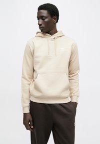 Bézs kapucnis pulóver elülső zsebbel és állítható húzózsinóros kapucnival. Fehér embléma a mellkason és bordázott mandzsetták jellemzik. Sötétbarna nadrággal párosítva.