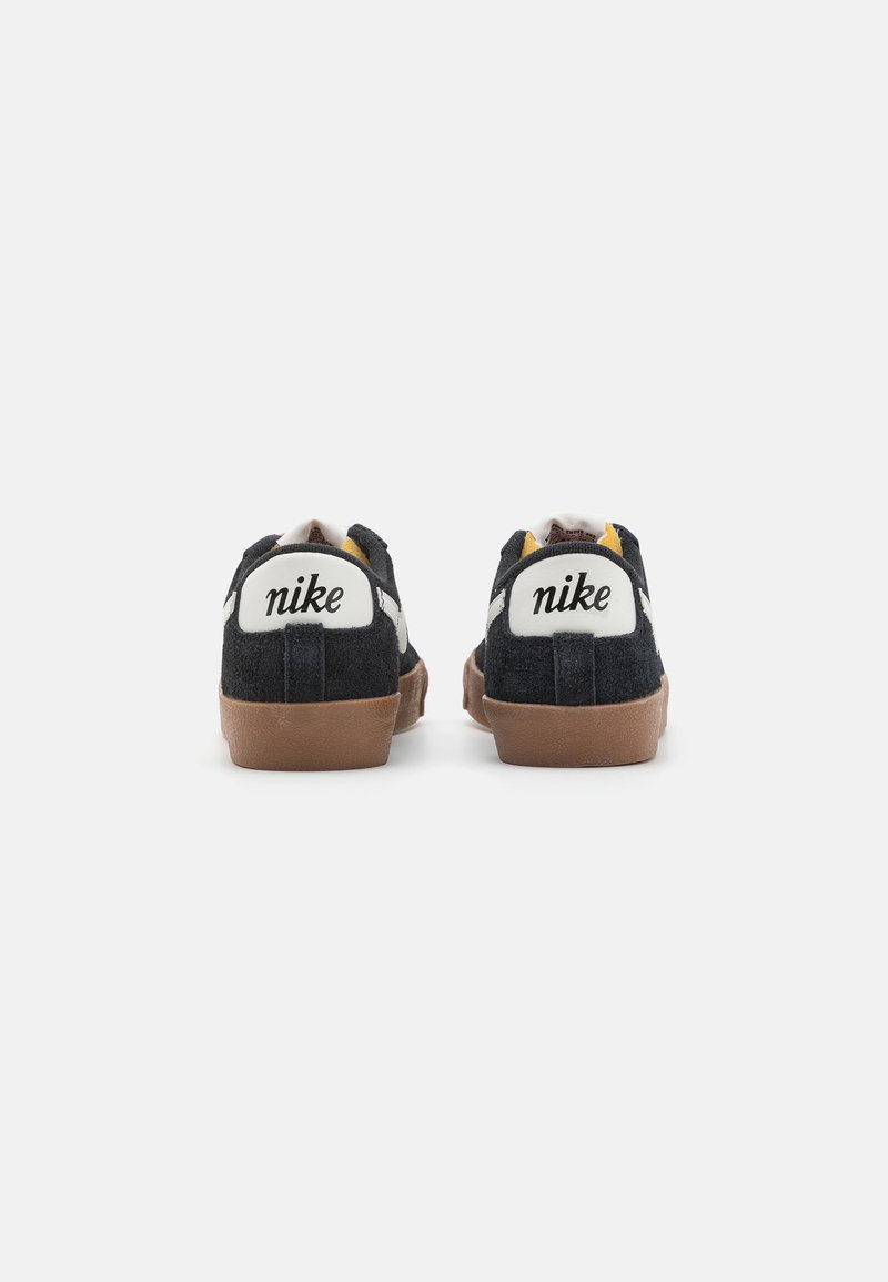 Zapatillas negras de ante con suela de goma, que presentan una marca blanca en el talón y acentos amarillos, mostrando un diseño de tipo bajo.