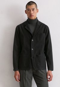 Dunkler Wollblazer mit zwei Vordertaschen, Ein-Knopf-Verschluss, spitzem Revers und strukturierter Oberfläche, getragen über einem passenden Rollkragenpullover.