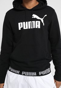 Sudadera negra de tela suave con un bolsillo tipo canguro en la parte frontal. Presenta un logo blanco de Puma y una banda con la marca en el dobladillo. Diseño sencillo y funcional.