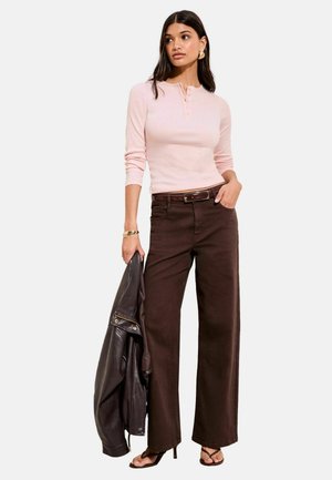 Svetloružový top s dlhým rukávom a gombíkmi, skombinovaný s tmavohnedými širokými nohavicami. Model drží čiernu koženú bundu. Lodičky zvýrazňujú outfit.