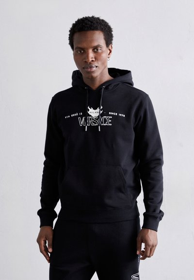 Schwarzer Baumwoll-Hoodie mit vorderer Kängurutasche, beinhaltet das weiße "Versace"-Logo und dekorative Elemente, verstellbare Kapuze mit Zugband, lässige Passform.