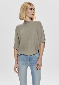 Khaki trui met een gestructureerd rib ontwerp, brede mouwen en een hoge halslijn, gecombineerd met lichtblauwe skinny jeans.