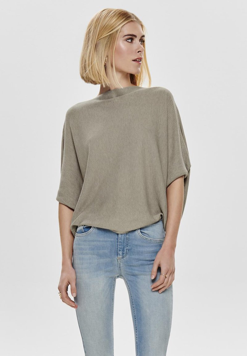 Khaki trui met een gestructureerd rib ontwerp, brede mouwen en een hoge halslijn, gecombineerd met lichtblauwe skinny jeans.