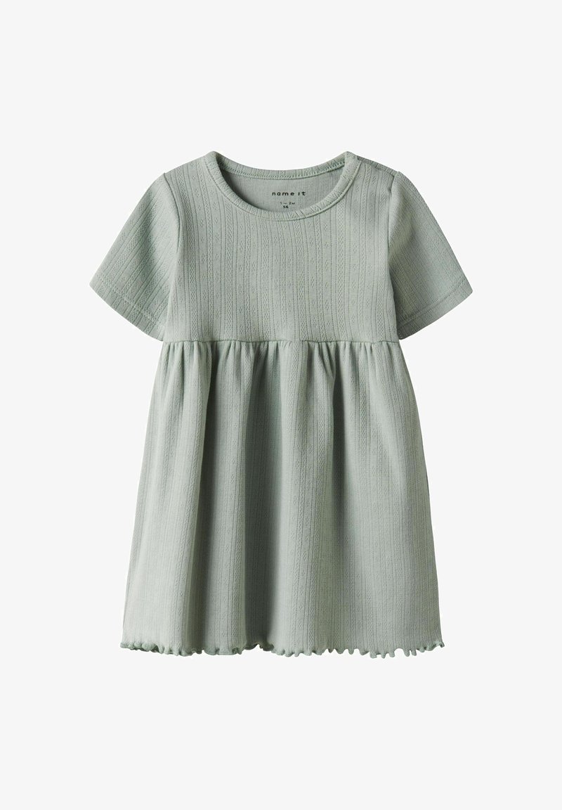 Šviesiai žalia suknelė su trumpomis rankovėmis, tekstūruotomis vertikaliomis juostelėmis, surinkta liemeniu ir banguotu apvadu. Pagaminta iš minkštos medvilnės ir sintetinio pluošto mišinio.